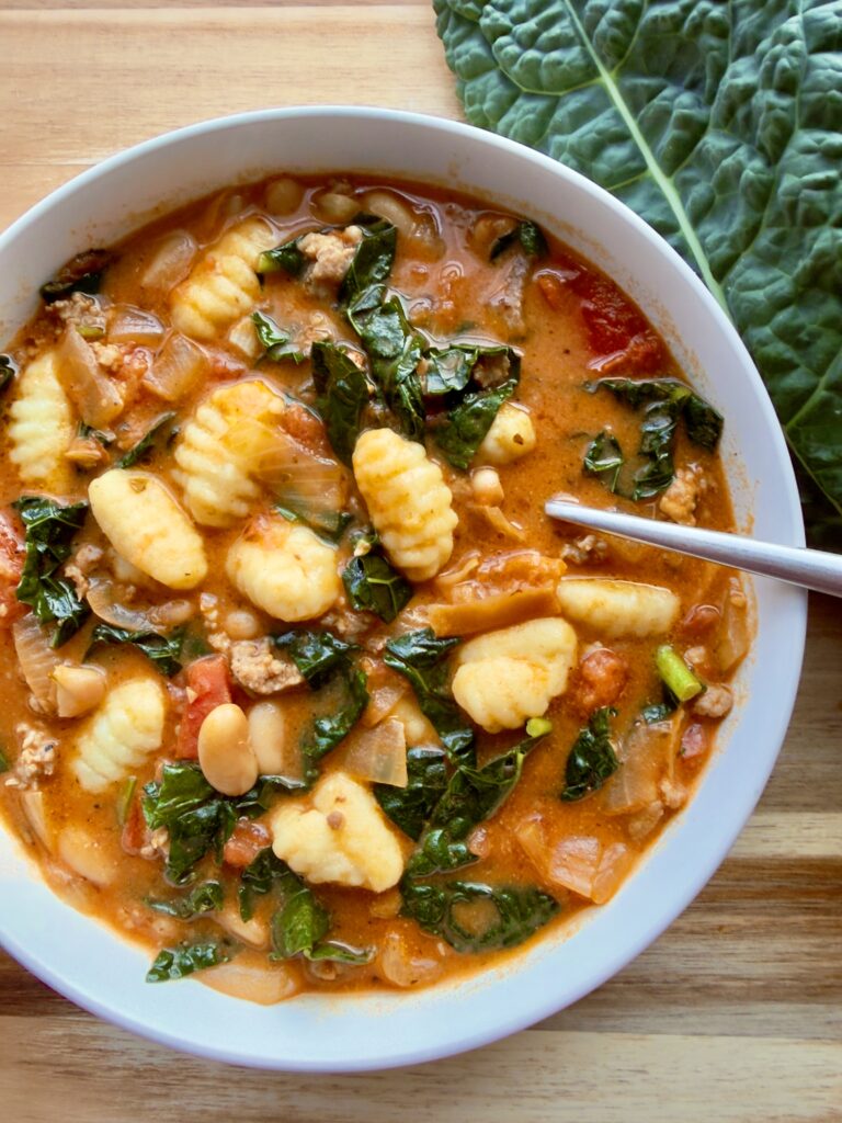 Creamy Tuscan sausage kale soup with gnocchi and white beans in a white bowl.