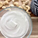 Creamy homemade cashew cream in a glass bowl surrounded by raw cashews on a wooden board.